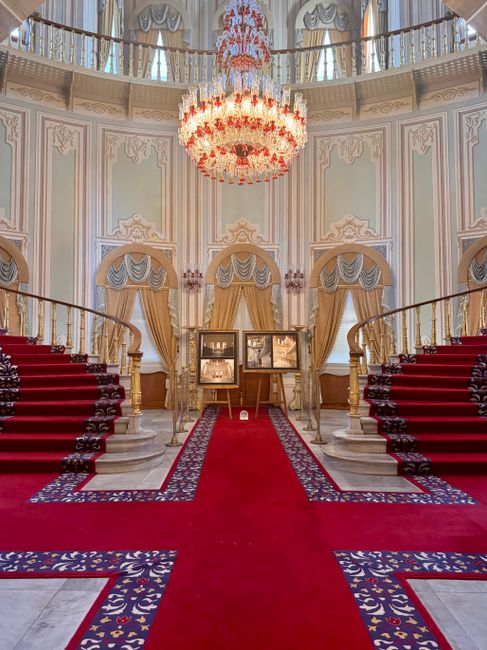 A grand interior staircase with red carpet leads down into a spacious, ornately decorated hall featuring cream marble columns with gold accents, intricate moldings, and a large archway beneath a statu