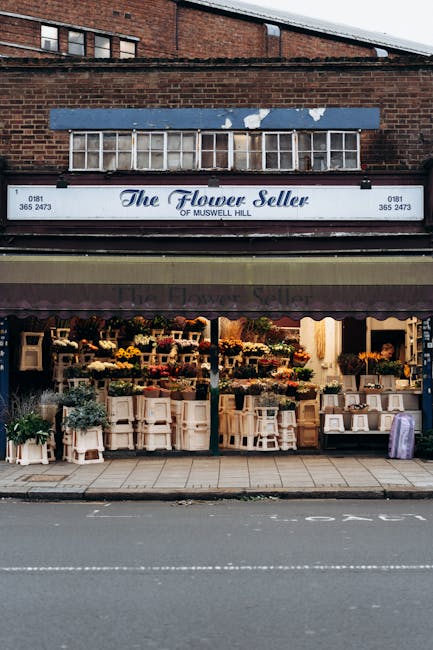 The exterior of a flower shop named 'The Flower Seller' located in Muswell Hill, with a blue signboard displaying the shop's name and contact information. The storefront features a maroon and beige aw