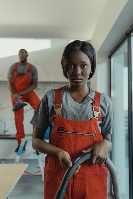 Three professional cleaners wearing orange uniforms are standing indoors, each holding a different cleaning tool. The central cleaner holds a vacuum cleaner with a white and grey cylindrical body, pos