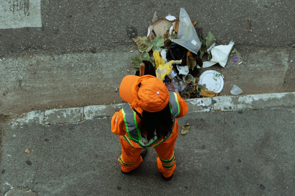 An overflowing waste disposal area in an urban setting, featuring a large metal recycling bin labeled for mixed paper and cardboard, with its lid open and various paper products spilling onto the surr